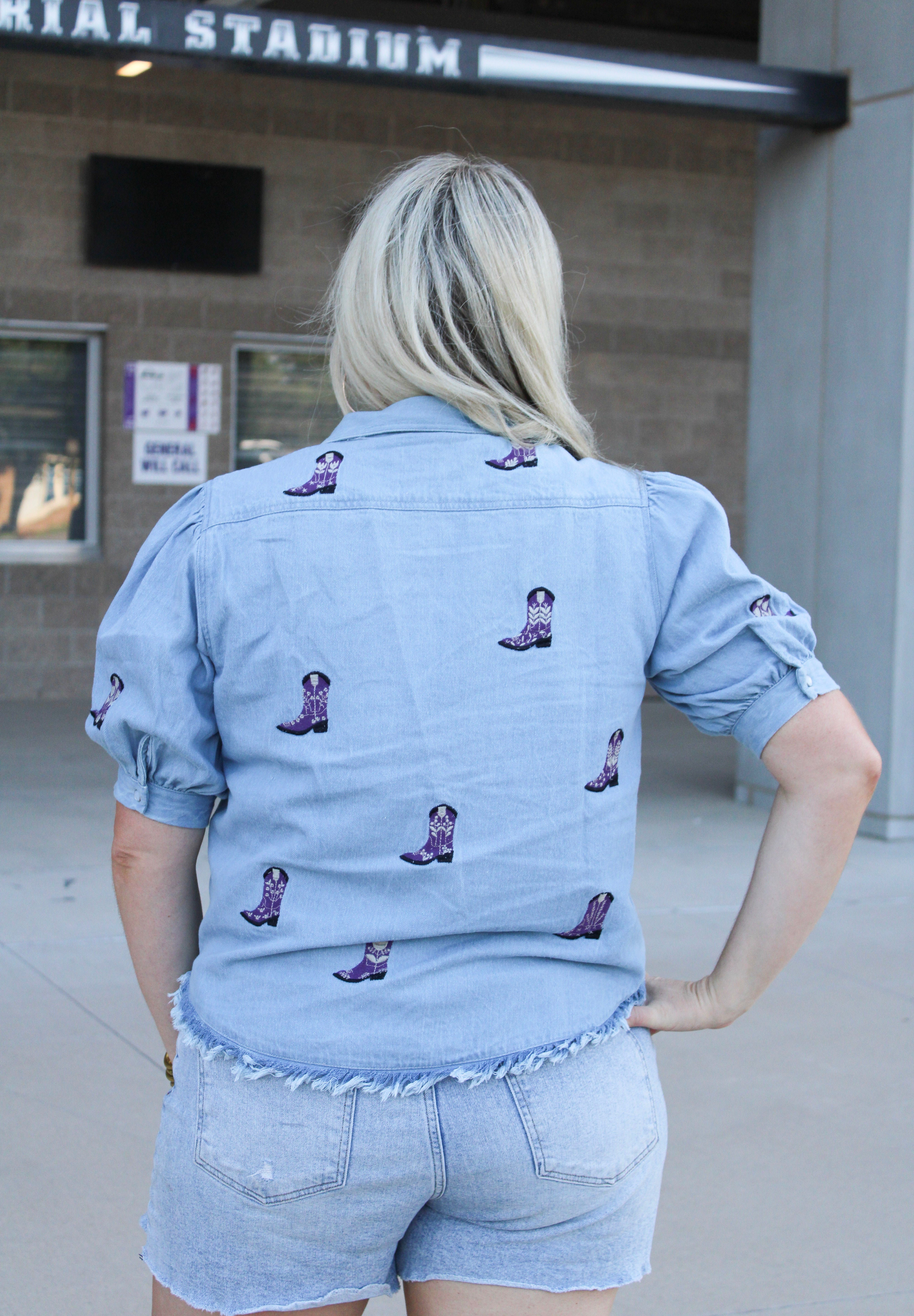 Woman with blonde hair standing in a back profile position wearing the Bailey Top in a lifestyle scene. The focal point of this image is the purple embroidered boots on back of top as well as back of ruffled sleeves. The model paired this top with light-wash denim shorts.