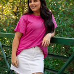 Woman with dark hair standing in a front profile position wearing the Basic Tee in a lifestyle scene. The focal point of this image is the bubbly pink colored tee. The model paired this tee with a white denim skirt.