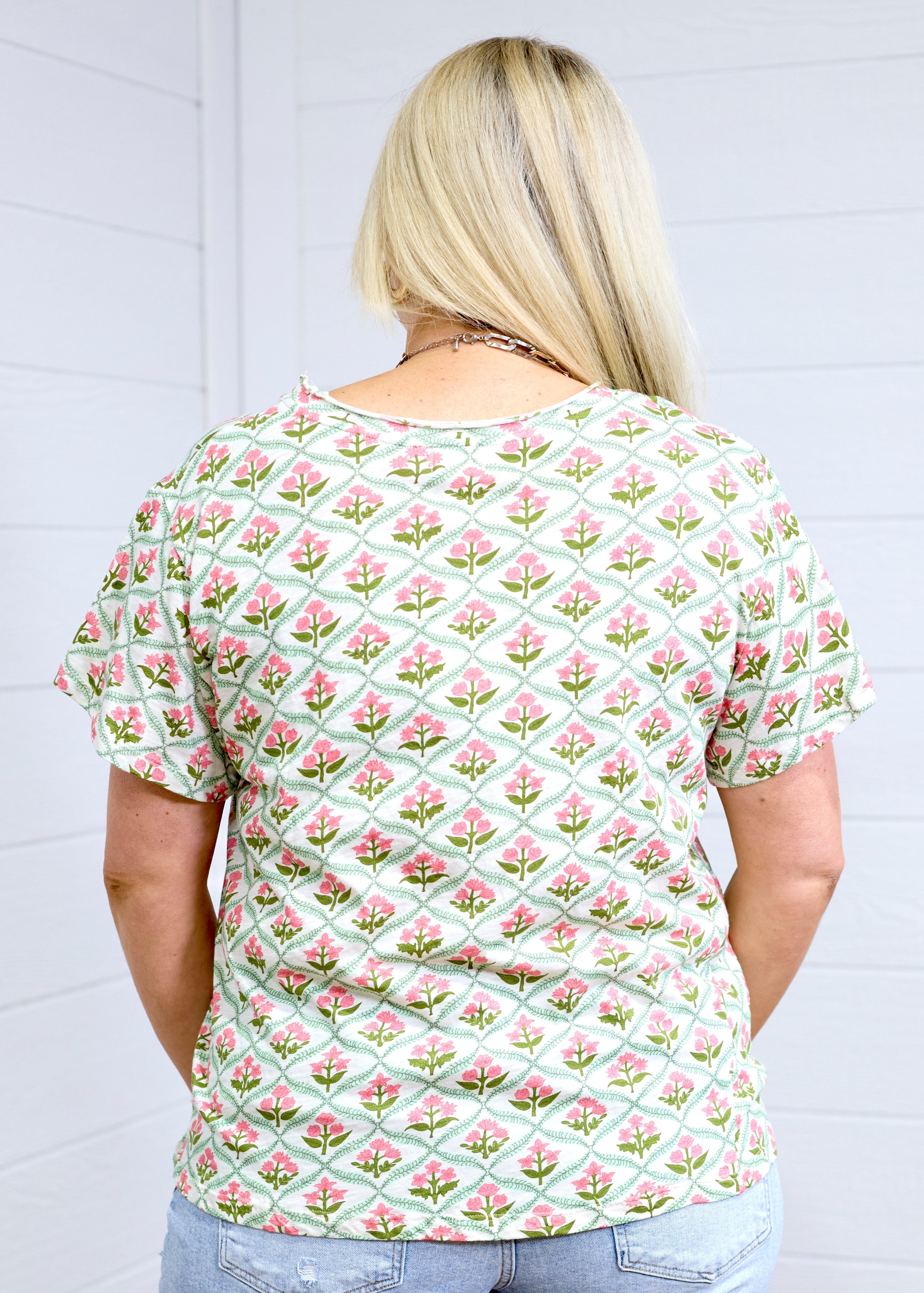 Woman with blonde hair standing in a back profile position wearing the Basic Tee against a white background. The focal point of this image is the pink and green floral pattern. The model paired this tee with light-wash denim shorts.