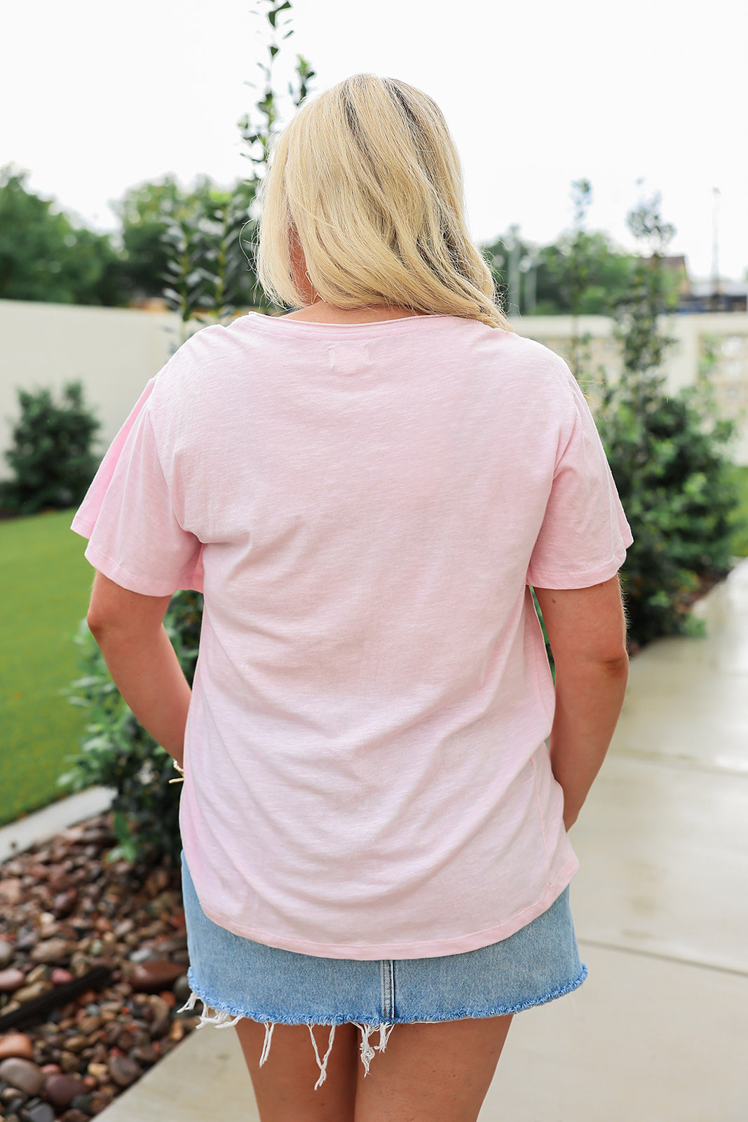 Woman with blonde hair standing in a back profile position wearing the Basic Tee in a lifestyle scene. The focal point of this image is the pink tee and length of tee. The model paired this tee with a light-wash denim skirt.