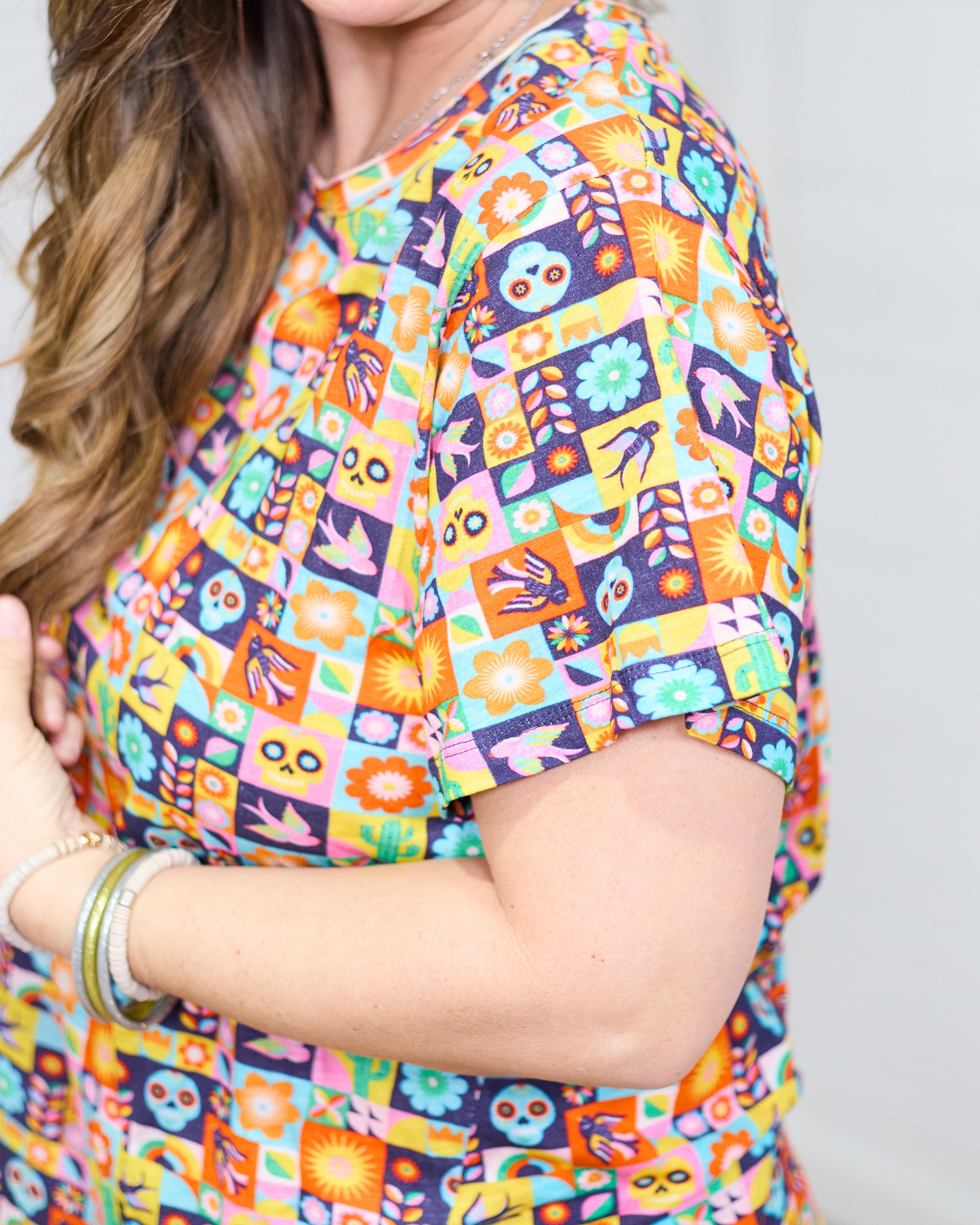 Woman with dark hair standing in a side profile position wearing the Basic Tee against a white background. The focal point of this image is the graphics on top (sugar scull, cacti, bird, etc...).