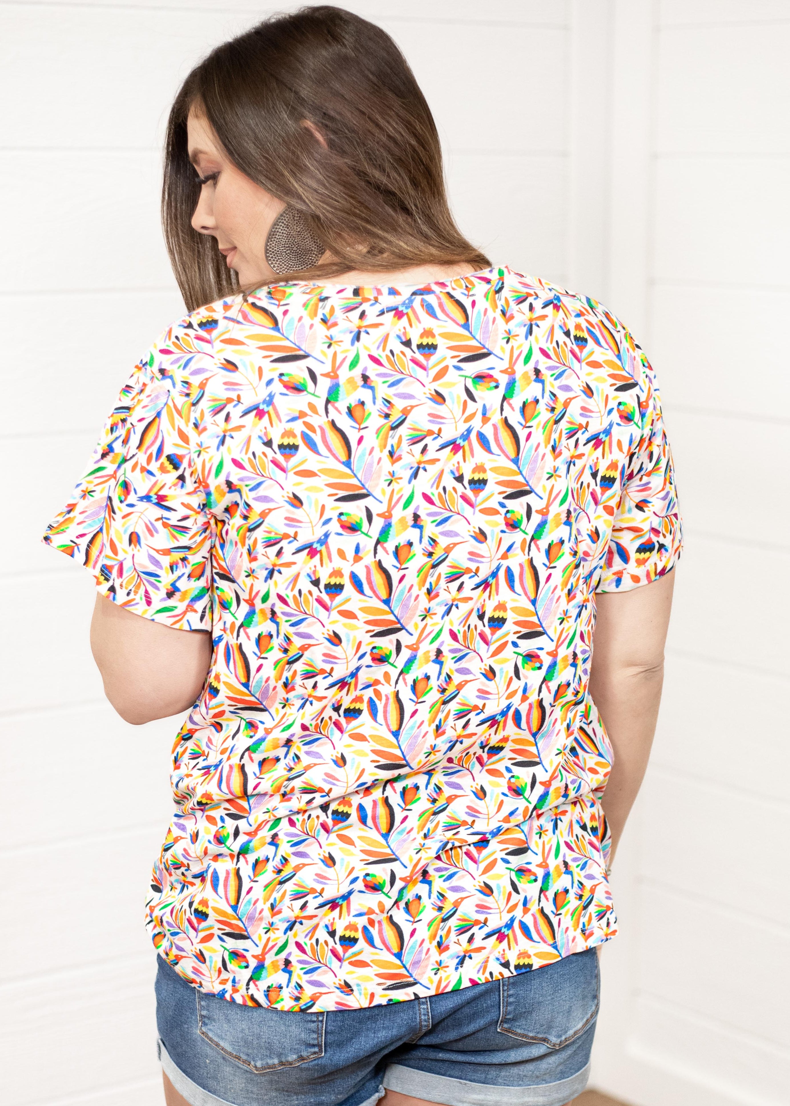 Woman with dark hair standing in a back profile position wearing the Basic Tee against a white background. The focal point of this image is colorful otomi graphics. The model paired this top with dark-wash denim shorts.
