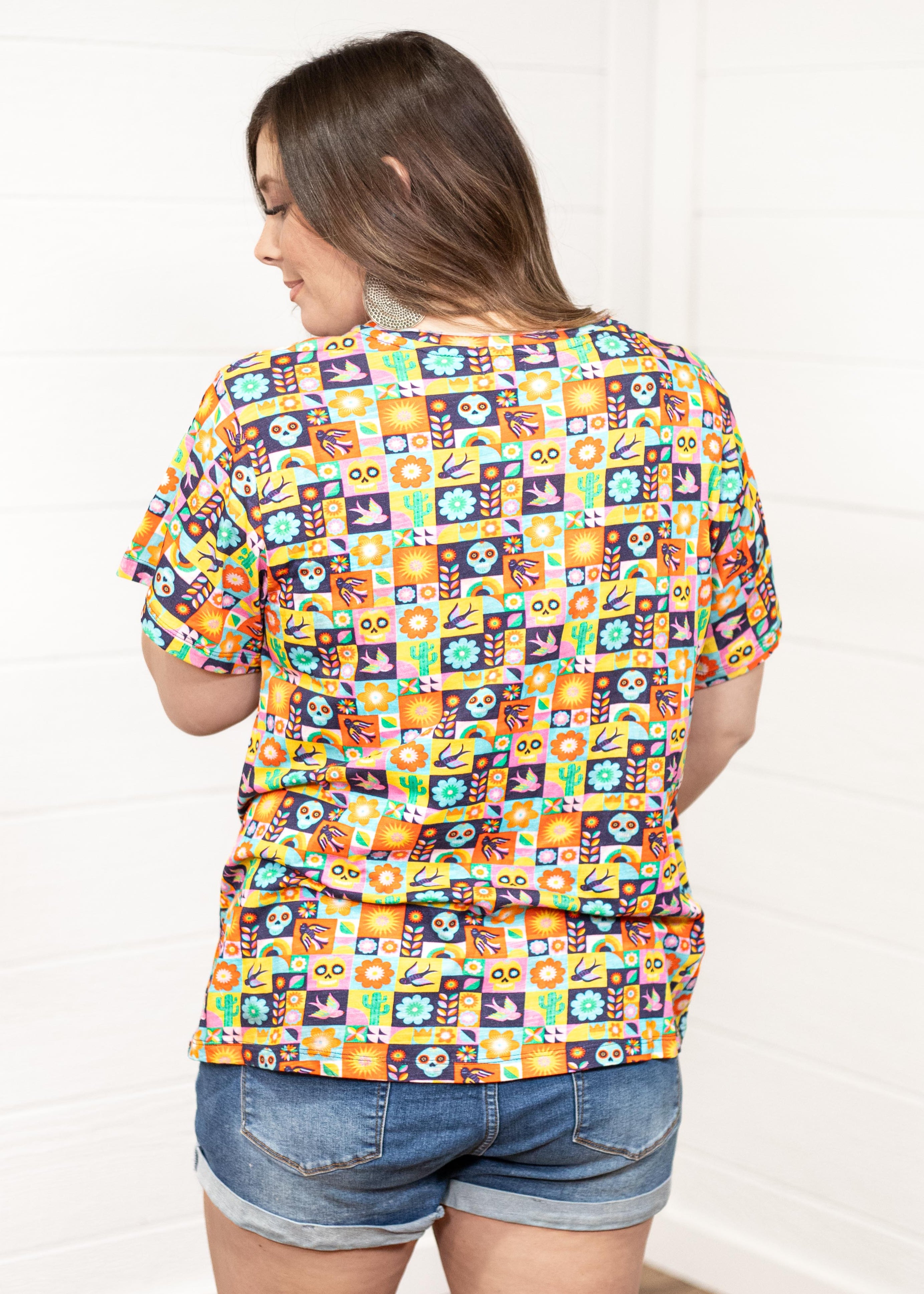 Woman with dark hair standing in a back profile position wearing the Basic Tee against a white background. The focal point of this image is the bright-colored graphics that include sugar skulls, cacti, etc. The model paired this tee with dark-wash denim shorts.