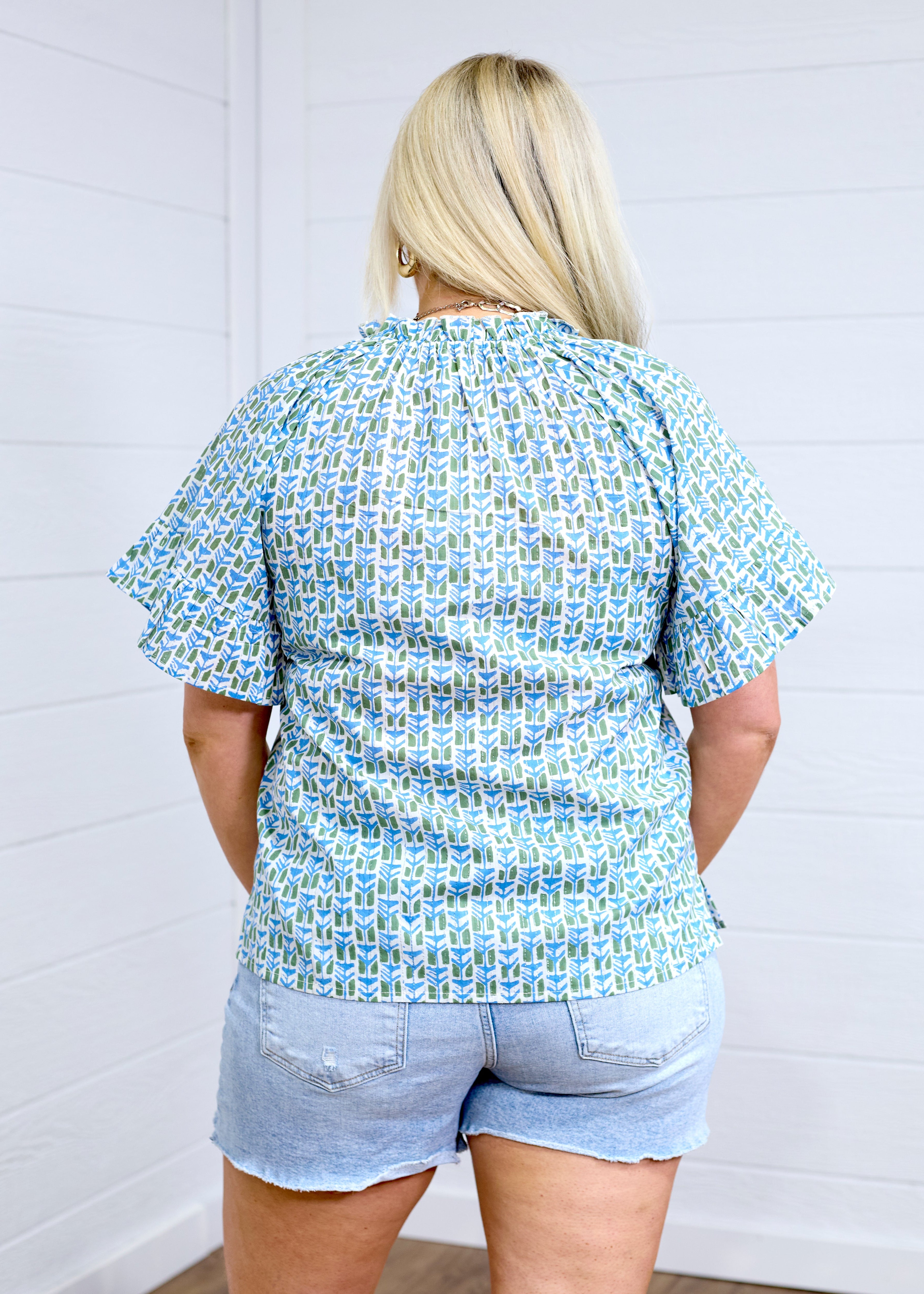 Woman with blonde hair standing in a back profile position wearing the Bell Top against a white background. The focal point of this image is the green and blue graphics, pleating on sleeves and top of collar. the model paired this top with light-wash denim shorts.
