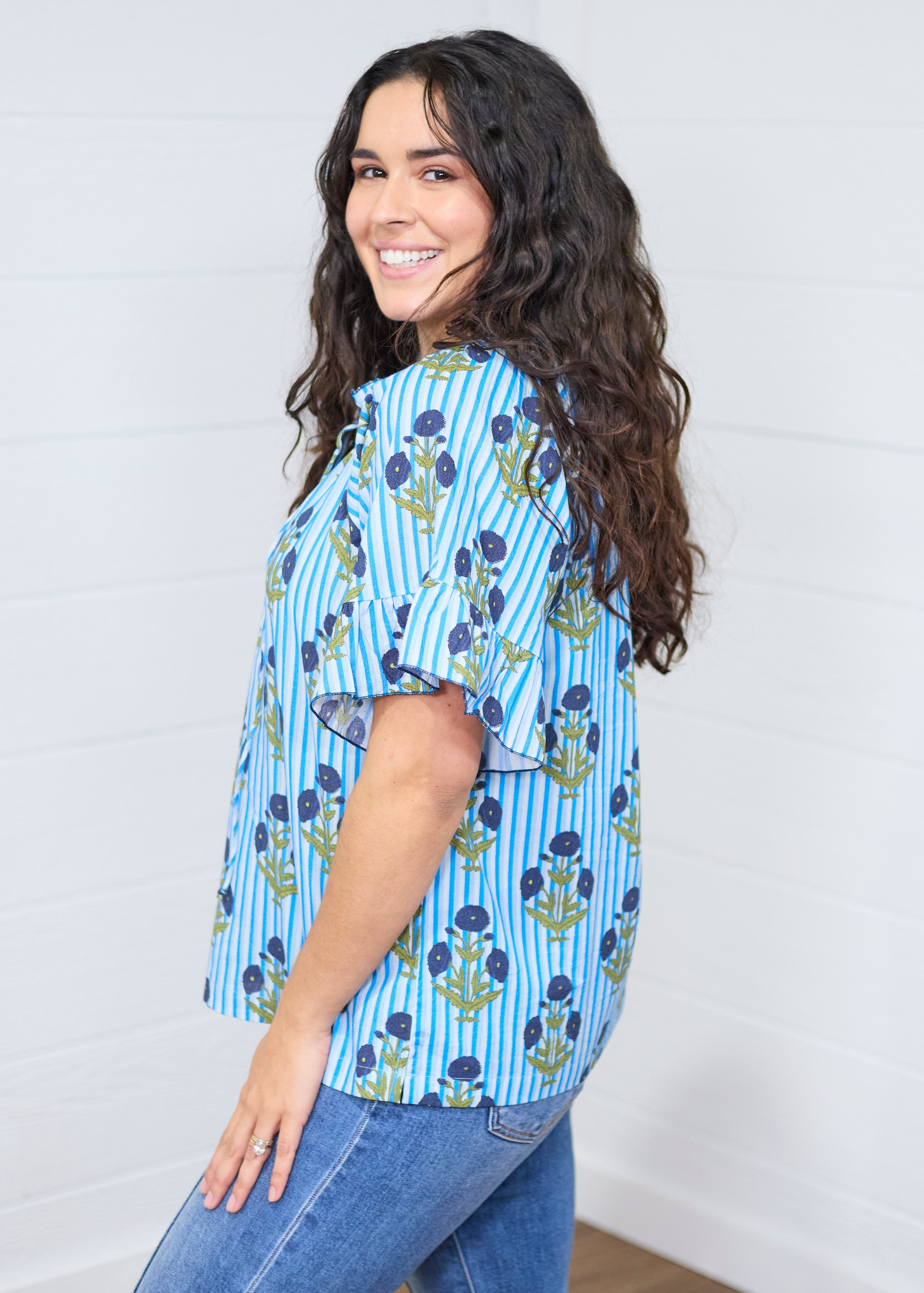 Woman with dark hair standing in a side profile position wearing the Bell Top against a white background. The focal point of this image is the graphics down the side of the top and the ruffle on the sleeve. The model paired this top with dark-wash denim jeans.