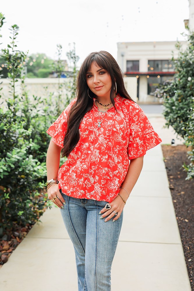 Woman with dark hair standing in a front profile position wearing the Bell Top in a lifestyle scene. The focal point of this image is the floral, tassels, and v-neckline. The model paired this top with light-wash denim jeans.