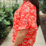 Woman with dark hair standing in a side profile position wearing the Bell Top in a lifestyle scene. The focal point of this image is the ruffled sleeves and white floral on red fabric. The model paired this top with light-wash denim jeans.