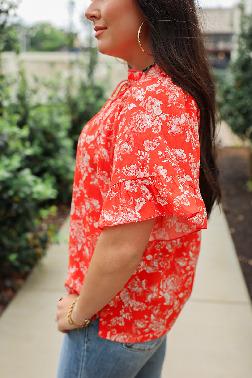 Woman with dark hair standing in a side profile position wearing the Bell Top in a lifestyle scene. The focal point of this image is the ruffled sleeves and white floral on red fabric. The model paired this top with light-wash denim jeans.