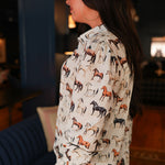 Woman with dark hair standing in a side profile position wearing the Caroline Top in a lifestyle scene. The focal point of this image is ruffle detail on cuff of sleeve and multi-color horse graphics. The model paired this top with dark-wash denim jeans.