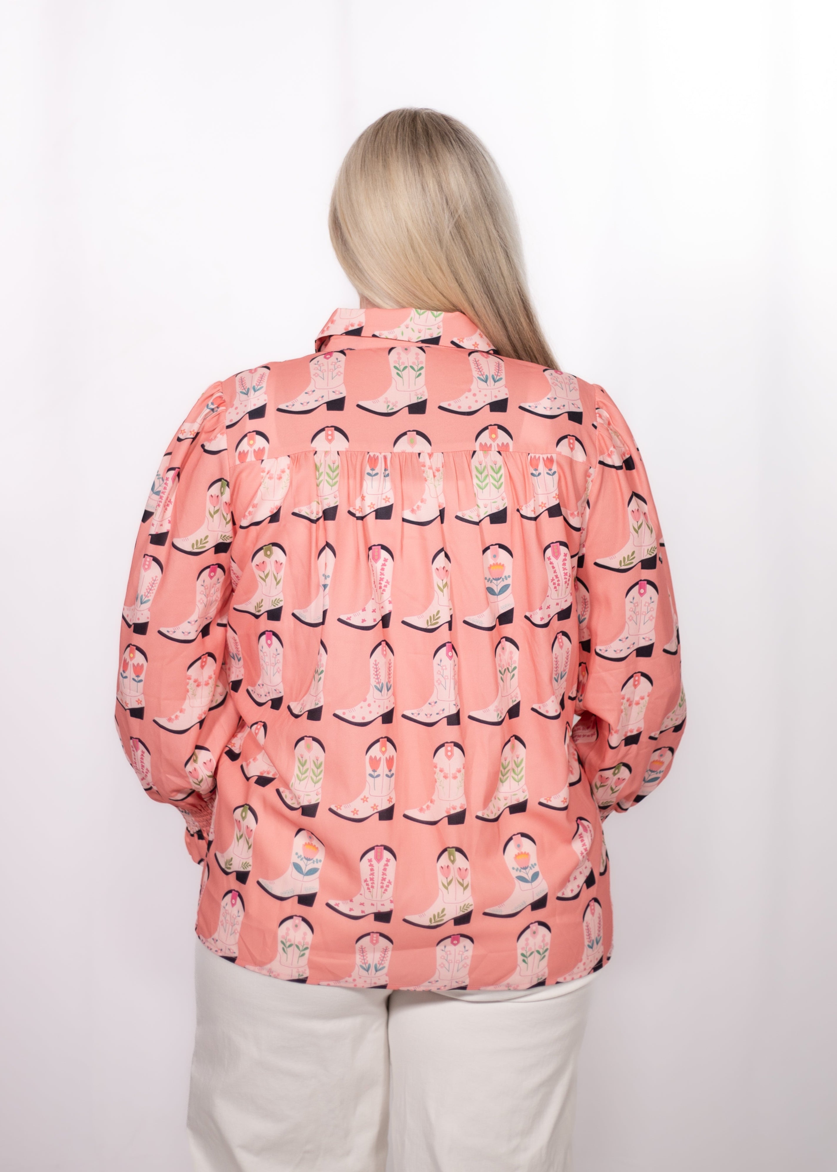 Woman with blonde hair standing in a back profile position wearing the Caroline Top against a white background. The focal point of this image is the boot graphics against pink shirt. The model paired this top with white pants.