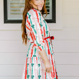 Woman with red hair wearing a long-sleeve dress with pink, red, and green graphic bows. Dress comes with red-fabric that is tied in front.