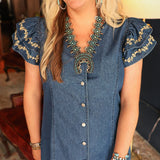 Woman with blonde hair standing in a front profile position wearing the Caylee Dress in a lifestyle scene. The focal point of this image is the buttons down front of dress, ruffled sleeves, and cream floral embroidery.