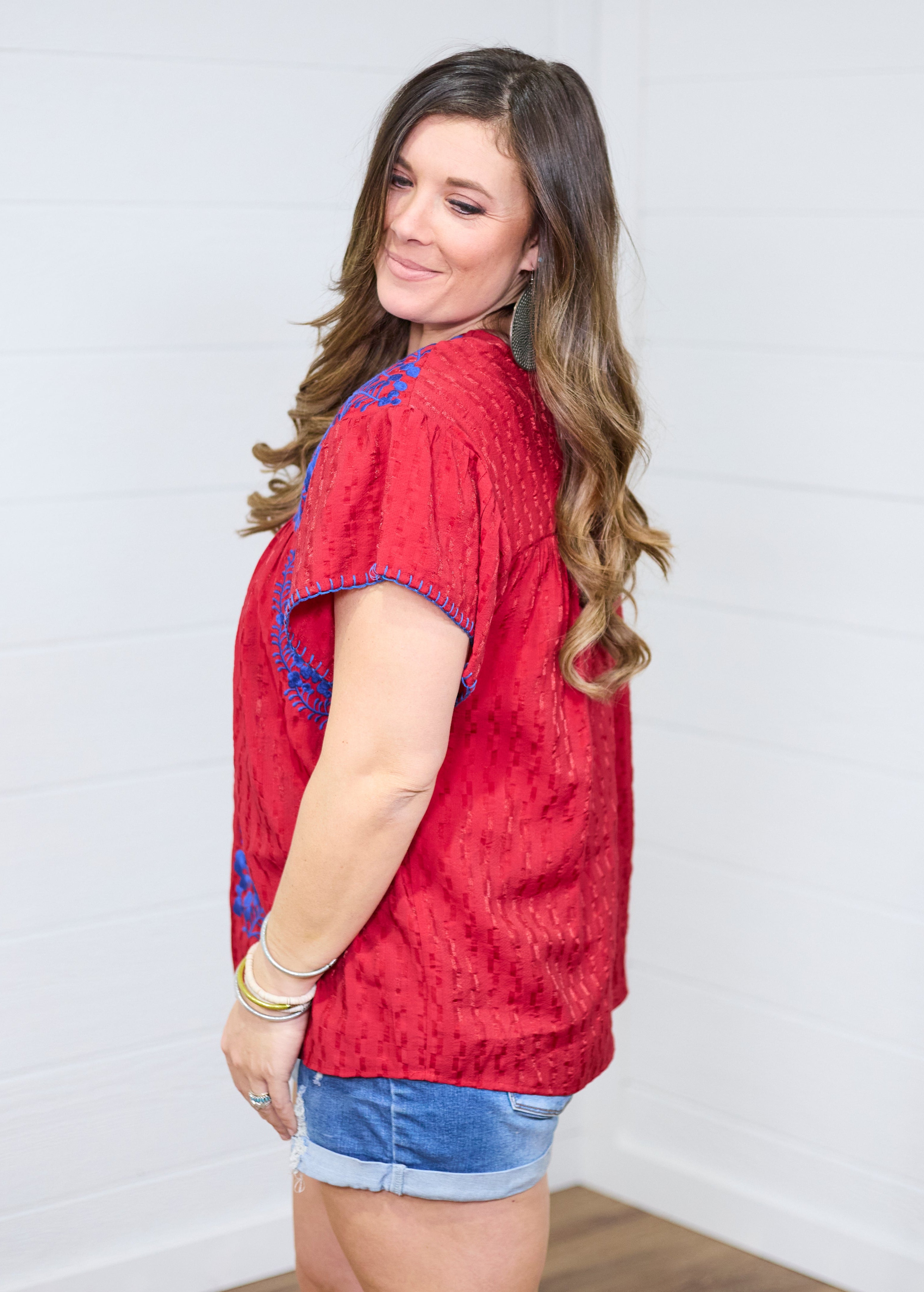 Woman with dark hair standing in a side profile position wearing the Chelsea Top against a white background. The focal point of this image is the blue embroidery on shoulders, loose sleeves, buck-stitch on cuff. The model paired this top with dark-wash denim shorts.