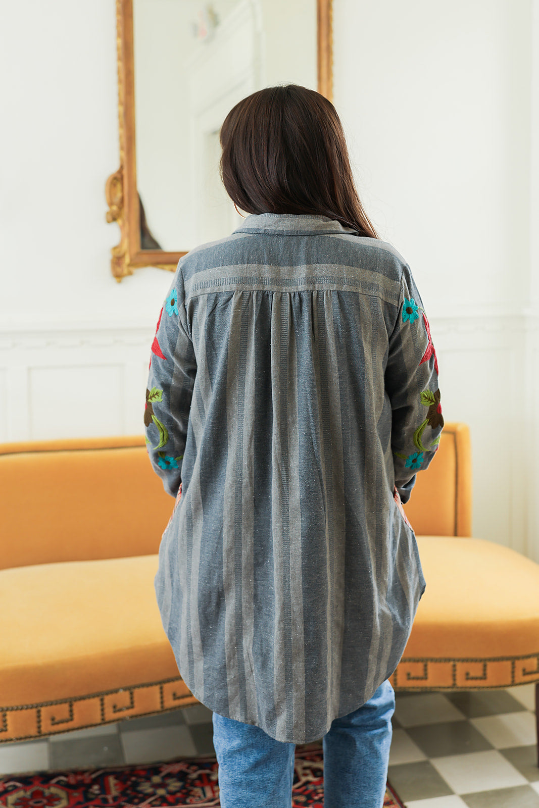 Woman with dark hair standing in a back profile position wearing the Diane Top in a lifestyle scene. The focal point of this image is the denim fabric and sneak peek of floral embroidery on sleeve. The model paired this top with mid-wash denim jeans.
