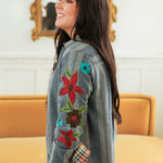 Woman with dark hair standing in a side profile position wearing the Diane Top in a lifestyle scene. The focal point of this image is the red, brown, green, and teal floral embroidery on sleeve. The model paired this top with mid-wash denim jeans.