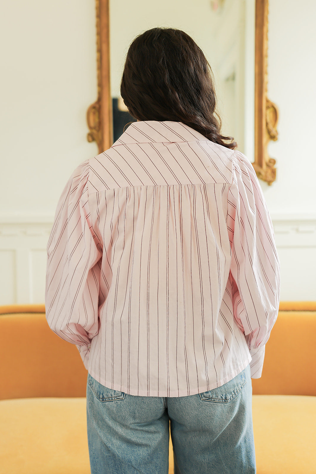 Woman with dark hair standing in a back profile position wearing the Dolly Top in a lifestyle scene. The focal point of this image is the pink and brown striped pattern on back as well as long sleeves. The model paired this top with light-wash denim jeans.