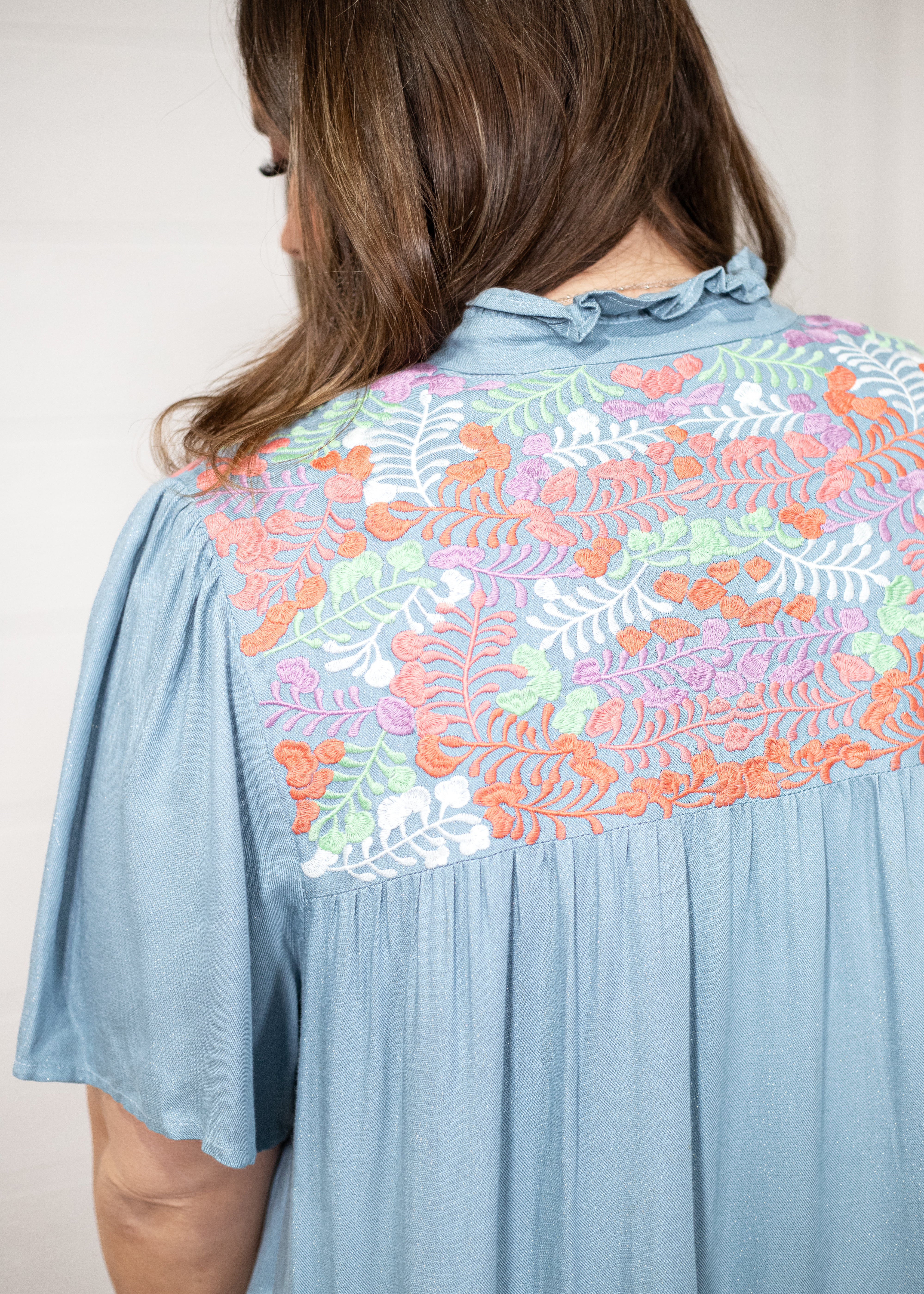 Woman with dark hair standing in a back profile position wearing the Evie Dress against a white background. The focal point of this image is floral pink, orange, white, and green embroidery.