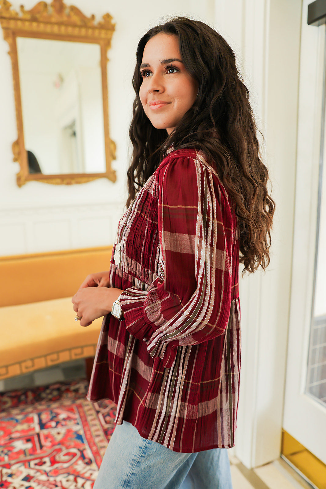 Woman with dark hair standing in a side profile position wearing the Faye Top in a lifestyle scene. The focal point of this image is the ruffled sleeve long sleeve as well as the length of top. The model paired this top with light-wash denim jeans.