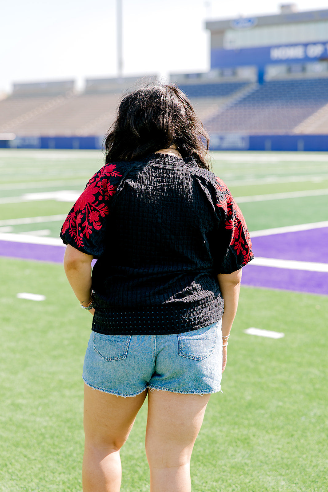 Woman with dark hair wearing a short-sleeve black top with red floral embroidery on sleeves. The model paired this top with light-wash denim shorts.