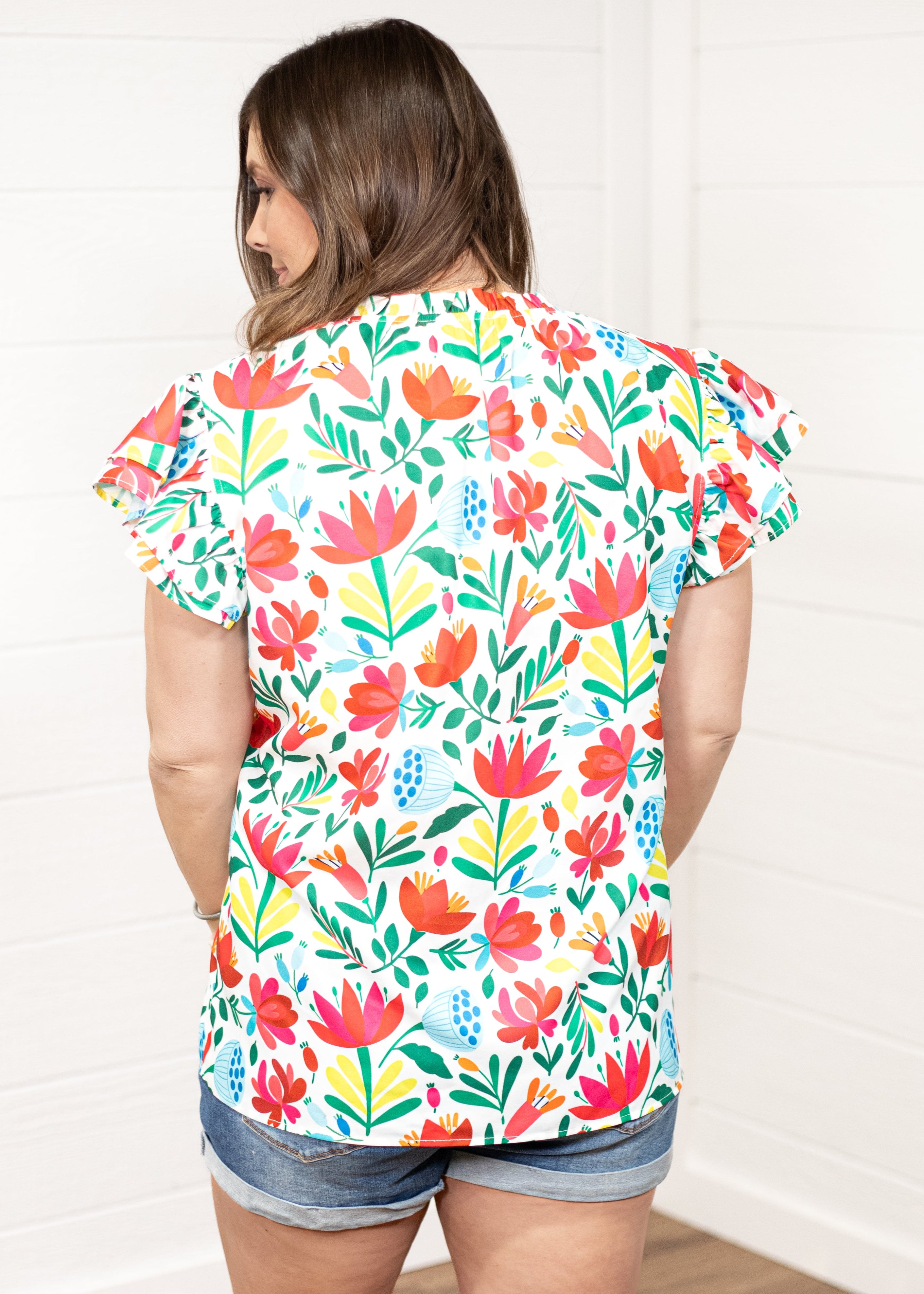 Woman with dark hair standing in a back profile position wearing the Iris Top against a white background. The focal point of this image is the colorful floral graphics on back of top. The model paired this top with dark-wash denim shorts.