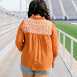 Woman with dark hair standing in a back profile position wearing the Jane Top at a football field. The focal point of this image is the white embroidery across shoulders as well as pleating beneath. The model paired this top with light-wash denim shorts.