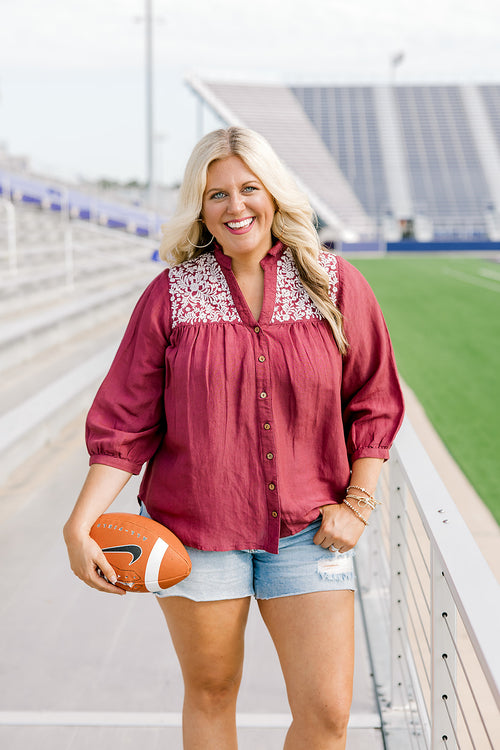 Woman with blonde hair standing in a front profile position wearing the Jane Top at a football field. The focal point of this image is the embroidery on the shoulders, buttons down the front of the top, and the v-neckline. The model paired this top with light-wash denim shorts.