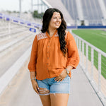 Woman with dark hair standing in a front profile position wearing the Jane Top at a football field. The focal point of this image is the buttons down the front of the top as well as the embroidery down shoulders and v-neckline. The model paired this top with light-wash denim shorts.