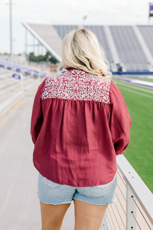 Woman with blonde hair standing in a back profile position wearing the Jane Top at a football field. The focal point of this image is the embroidery across the shoulders and pleating beneath. The model paired this top with light-wash denim shorts.