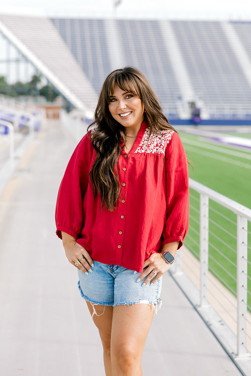 Woman with dark hair standing in a front profile position wearing the Jane Top at a football field. The focal point of this image is the buttons down the front of top, pleating beneath embroidery on shoulders, and quarter sleeves with pleating towards cuff. The model paired this top with light-wash denim shorts.