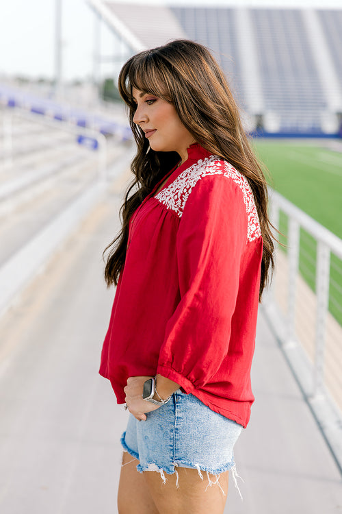 Woman with dark hair standing in a side profile position wearing the Jane Top at a football field. The focal point of this image is the quarter sleeve with pleating to the cuff as well as embroidery over the shoulders. The model paired this top with light-wash denim shorts.
