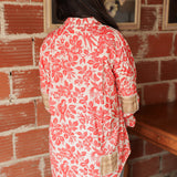 Woman with dark hair standing in a back profile position wearing the Karen Top against a red brick background. The focal point of this image is plaid patches on back and elbows of top as well as the red bird/floral graphics. The model paired this top with light-wash denim jeans.