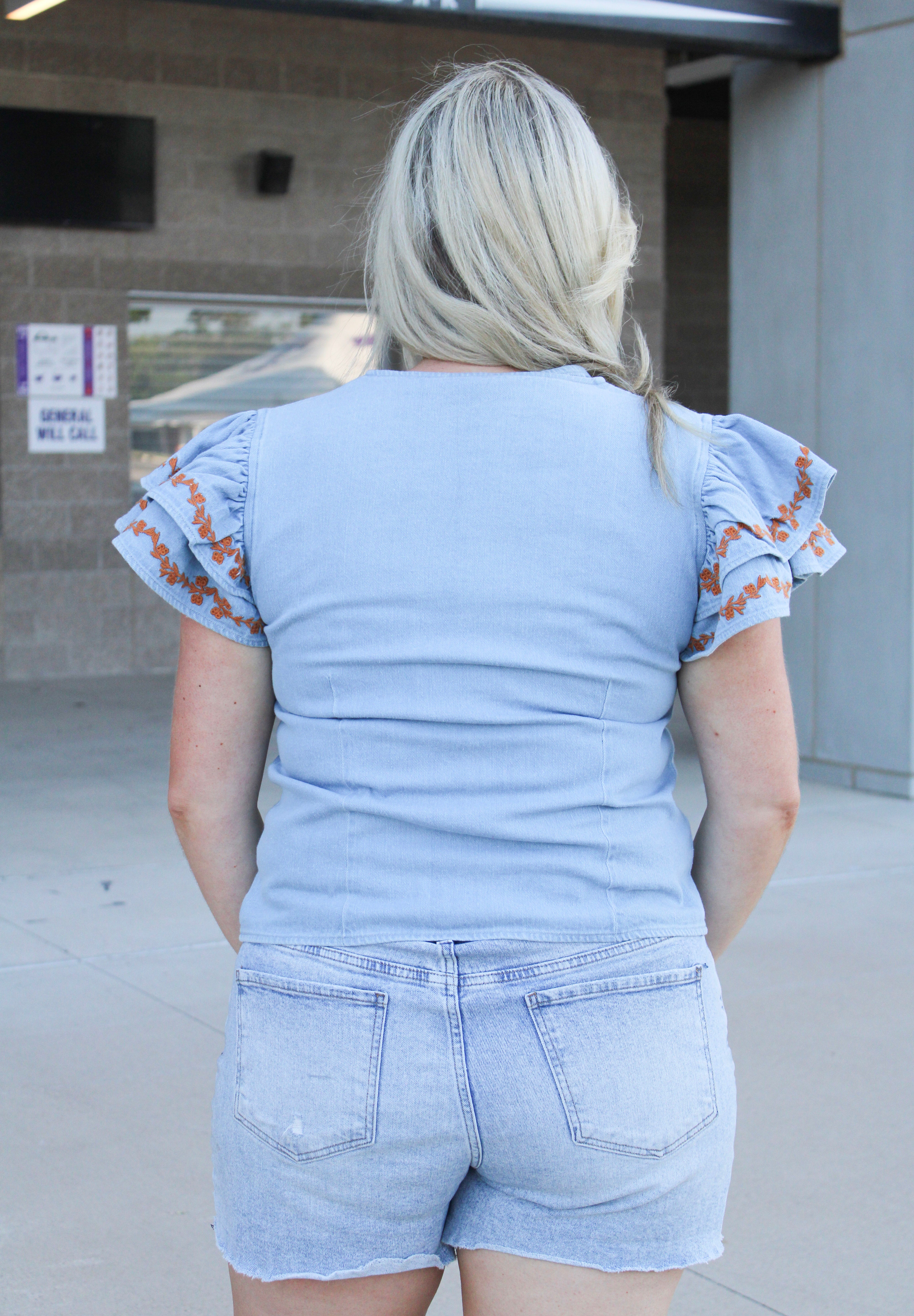 Woman with blonde hair wearing denim top with burnt orange embroidery on sleeves, and light-wash denim shorts.