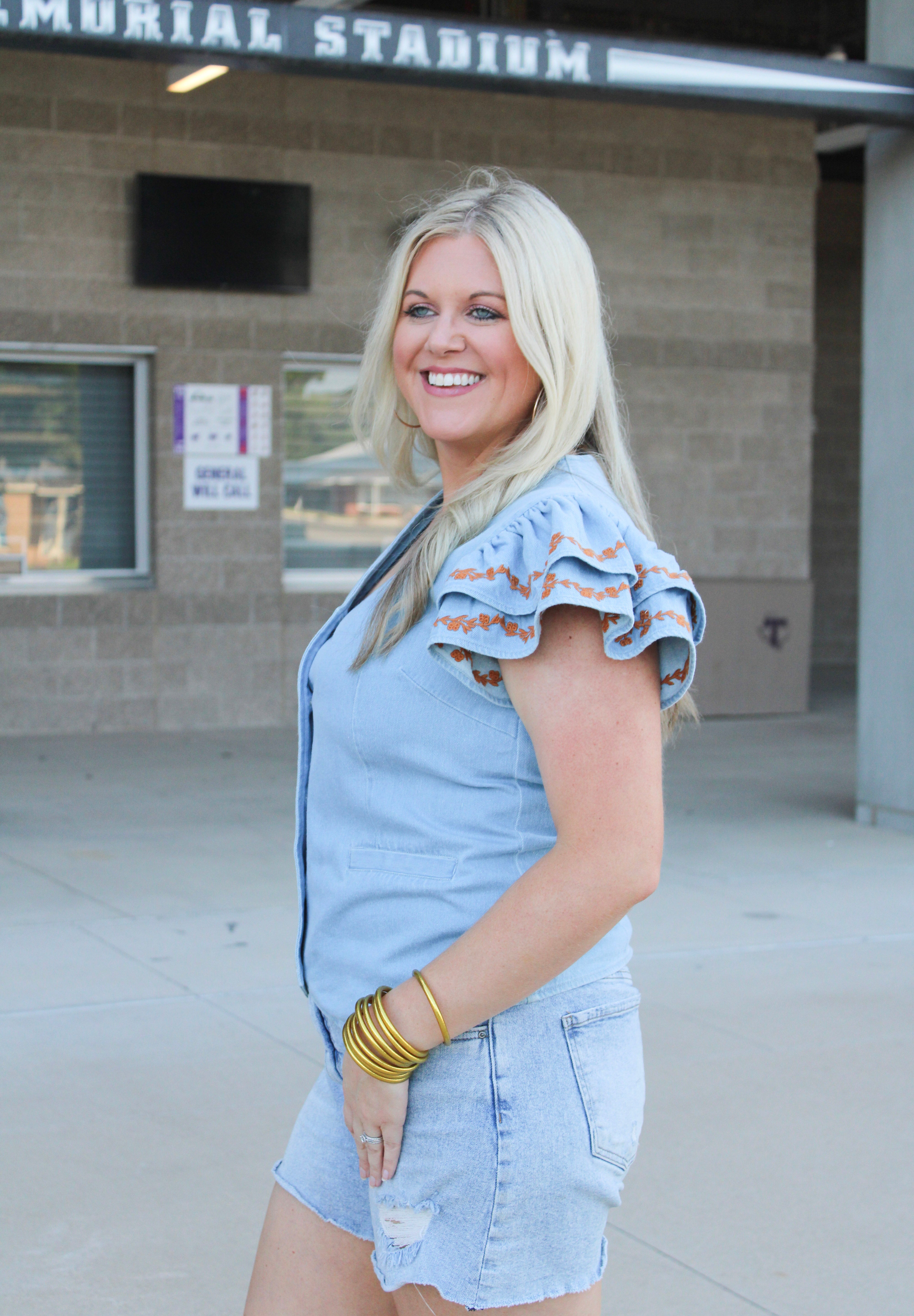 Woman with blonde hair wearing a denim top with burnt orange embroidery on sleeves, and light-wash denim shorts.
