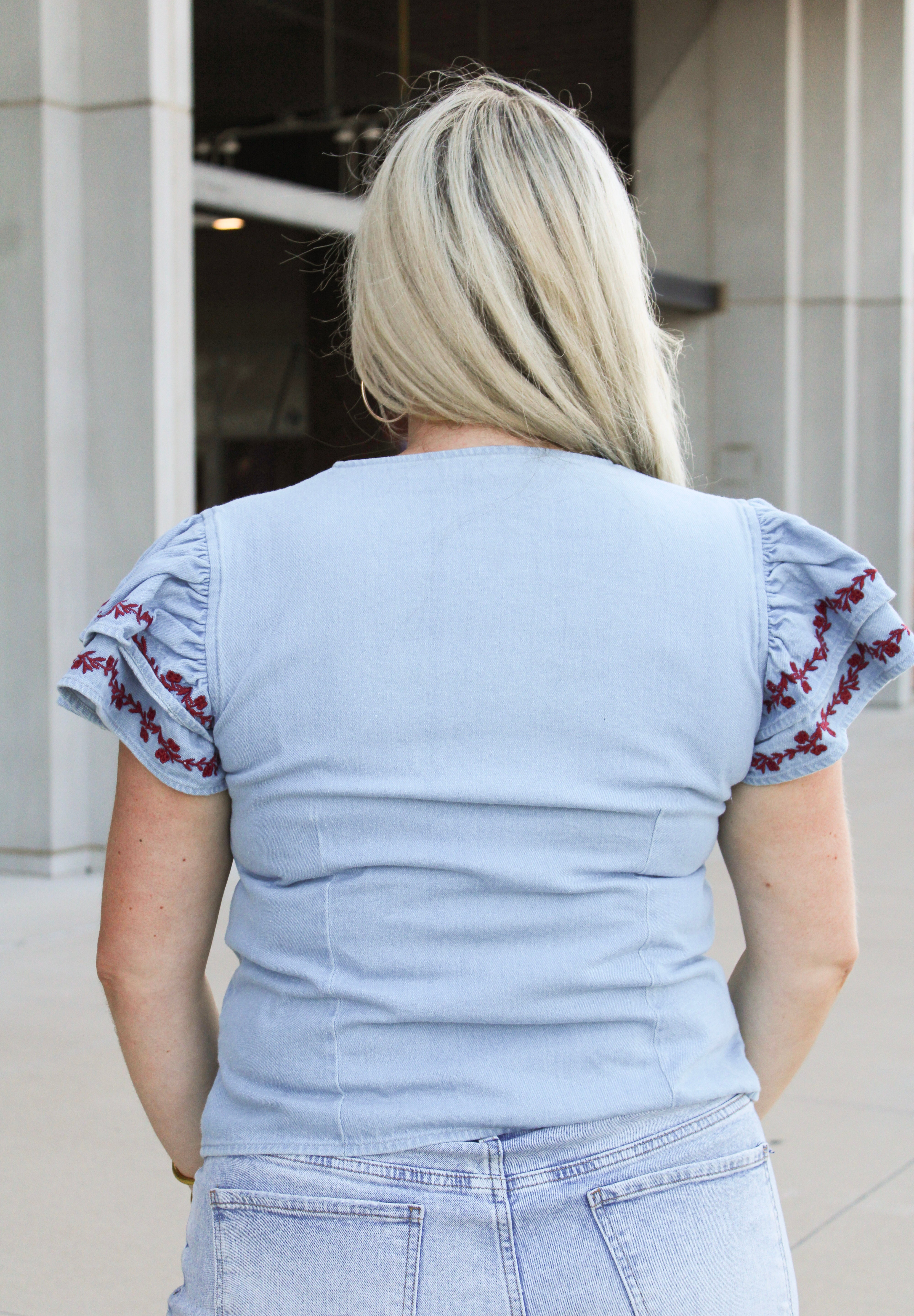 Woman with blonde hair wearing a short-sleeve denim top with maroon floral embroidery on sleeves, and light-wash denim shorts.
