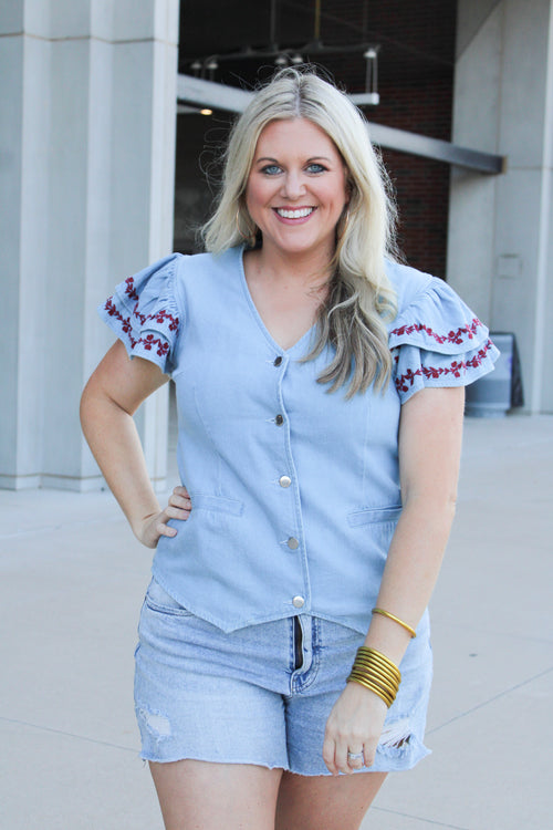 Woman with blonde hair wearing a short-sleeve denim top with maroon floral embroidery on sleeves, and light-wash denim shorts.
