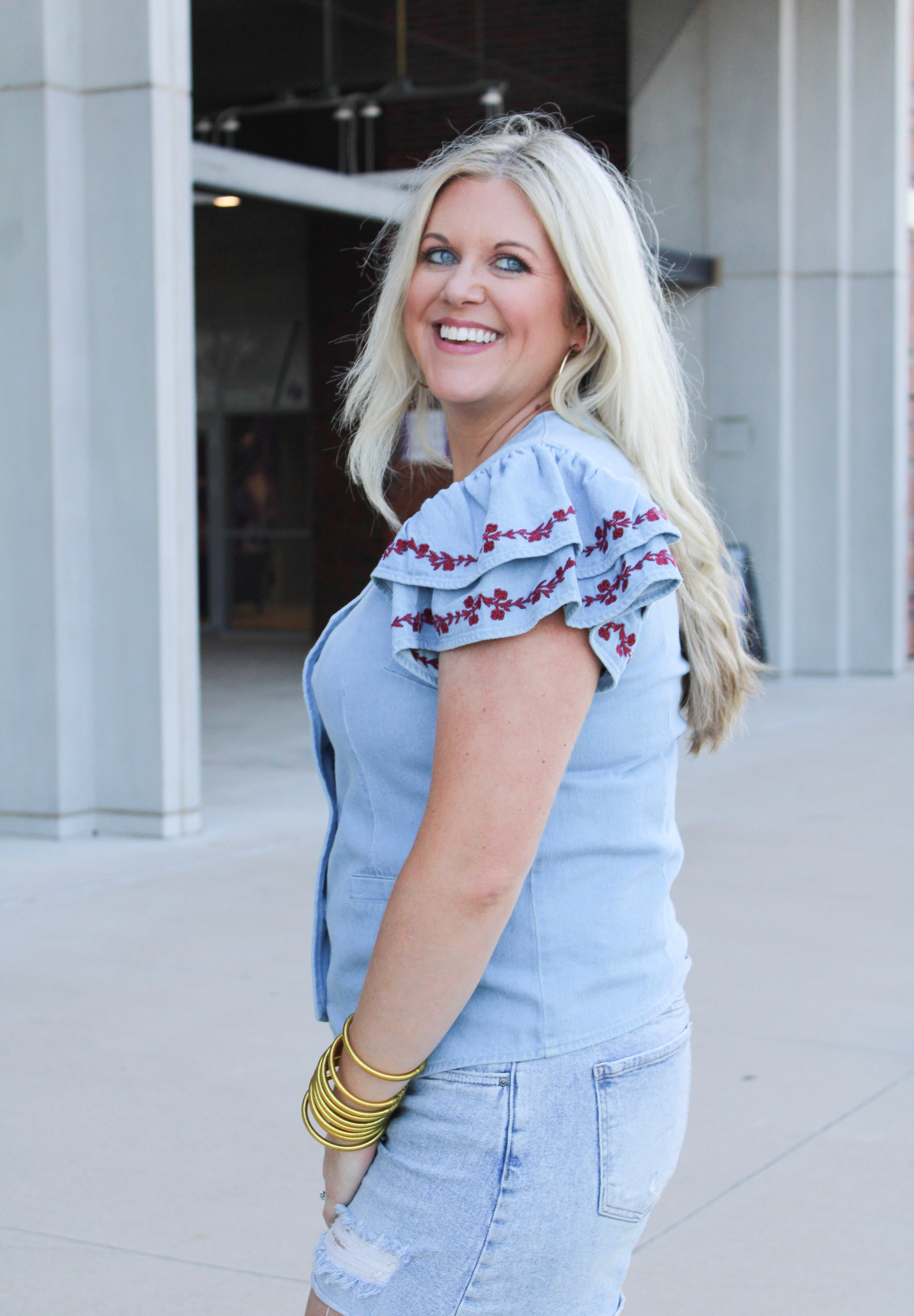 Woman with blonde hair wearing a short-sleeve denim top with maroon floral embroidery on sleeves, and light-wash denim shorts.