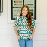Woman with red hair wearing a short-sleeve top with Christmas item graphics, ruffled/pleated sleeves, and ruffled collar. Model paired this top with denim jeans.