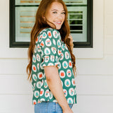 Woman with red hair wearing a short-sleeve top with Christmas item graphics, ruffled/pleated sleeves, and ruffled collar. Model paired this top with denim jeans.