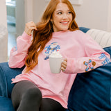 Girl sitting on couch in pink sweatshirt with appliqué Santa in blue & white on front. Christmas icons in blue & white down both sleeves.