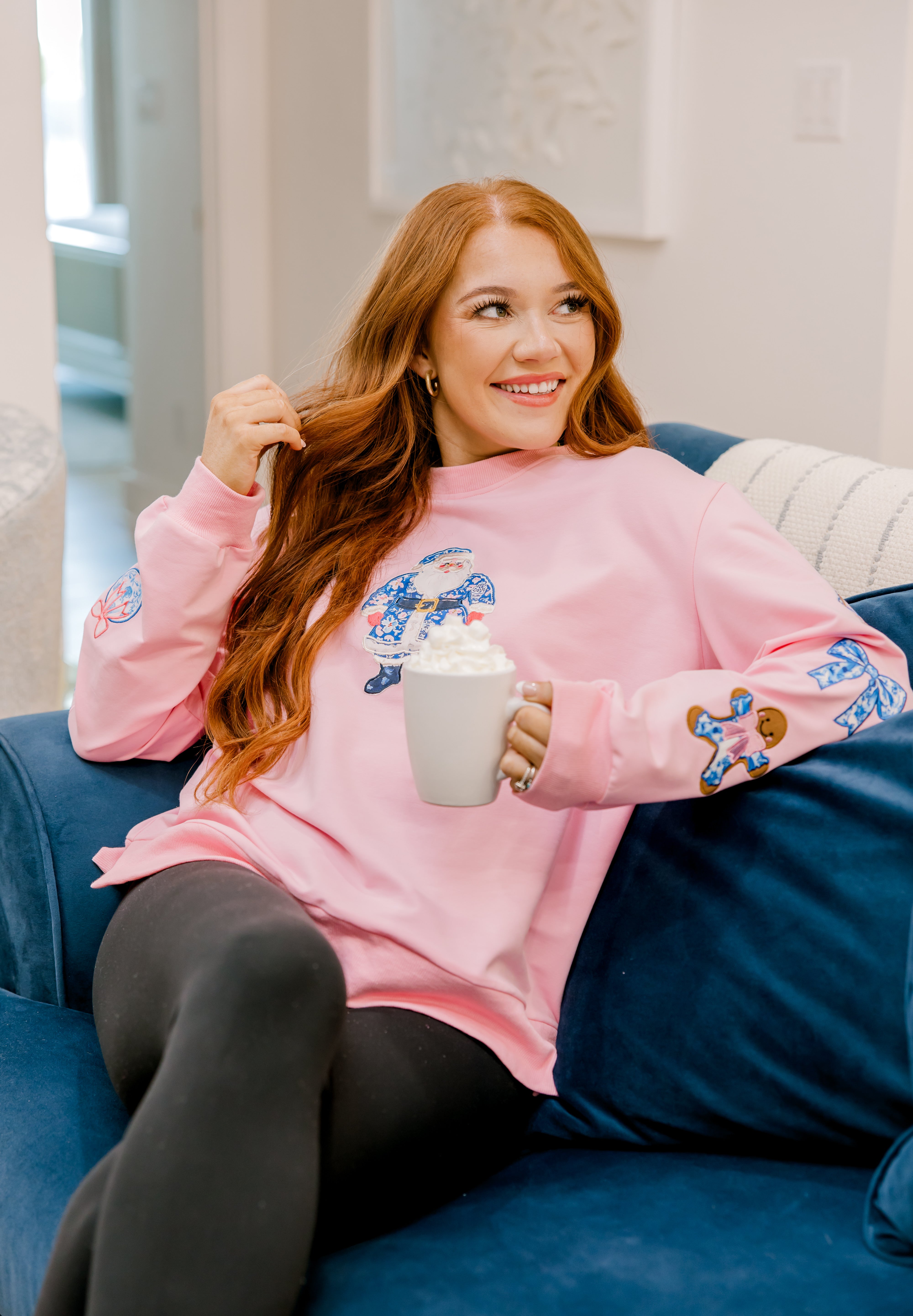 Girl sitting on couch in pink sweatshirt with appliqué Santa in blue & white on front. Christmas icons in blue & white down both sleeves.