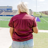 Woman with blonde hair standing in a back profile position wearing the Lilly Top in a lifestyle scene. The focal point of this image is the maroon eyelet pattern as well as pleating on back and ruffle on collar. The model paired this top with light-wash denim shorts.