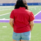 Woman facing backwards wearing a short-sleeve red eyelet top and light-wash denims shorts.