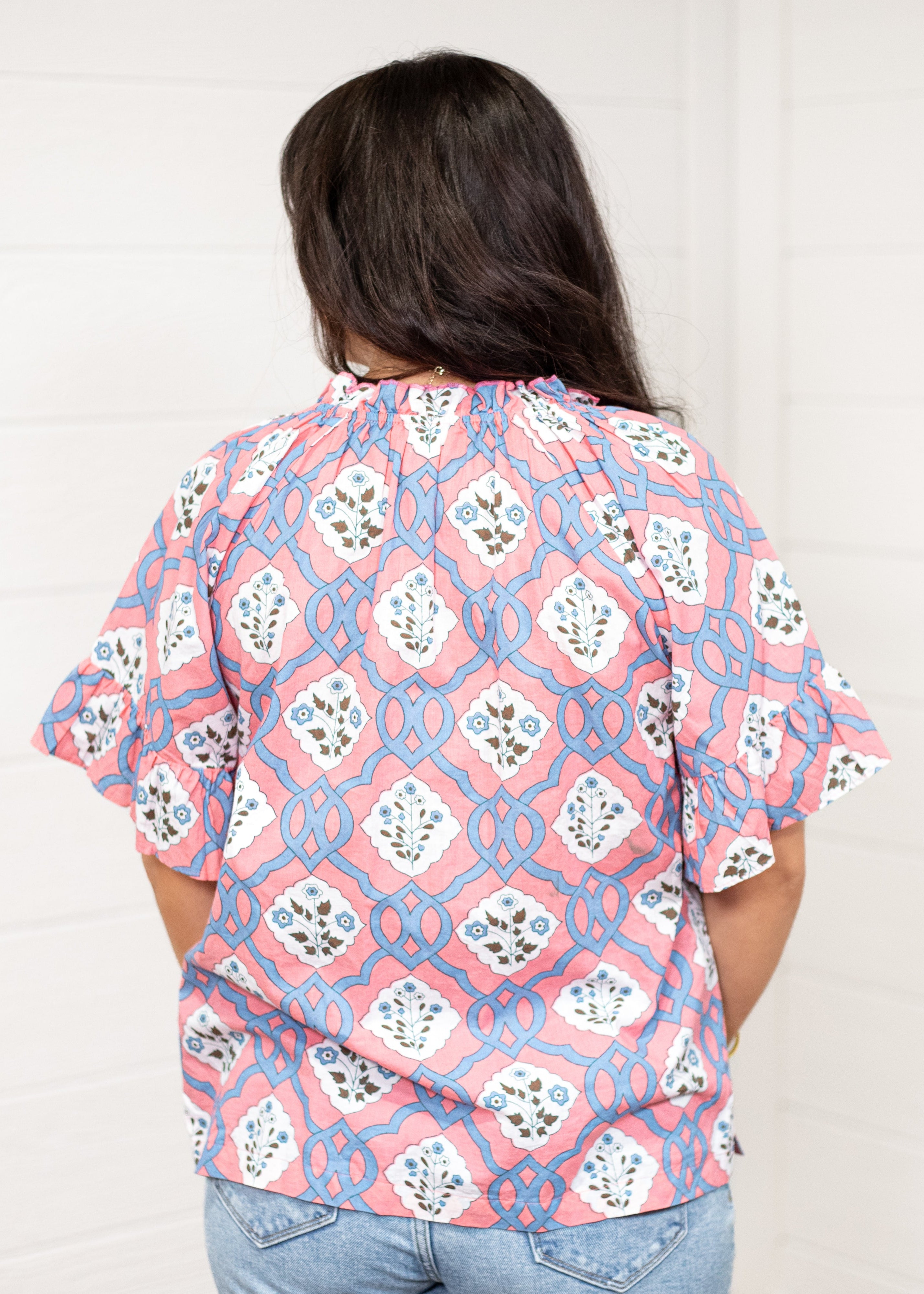 Woman with dark hair standing in a back profile position wearing the Mia Top against a white background. The focal point of this image is the intricate blue and pink floral design as well as pleating on collar and sleeves. The model paired this top with light-wash denim jeans.
