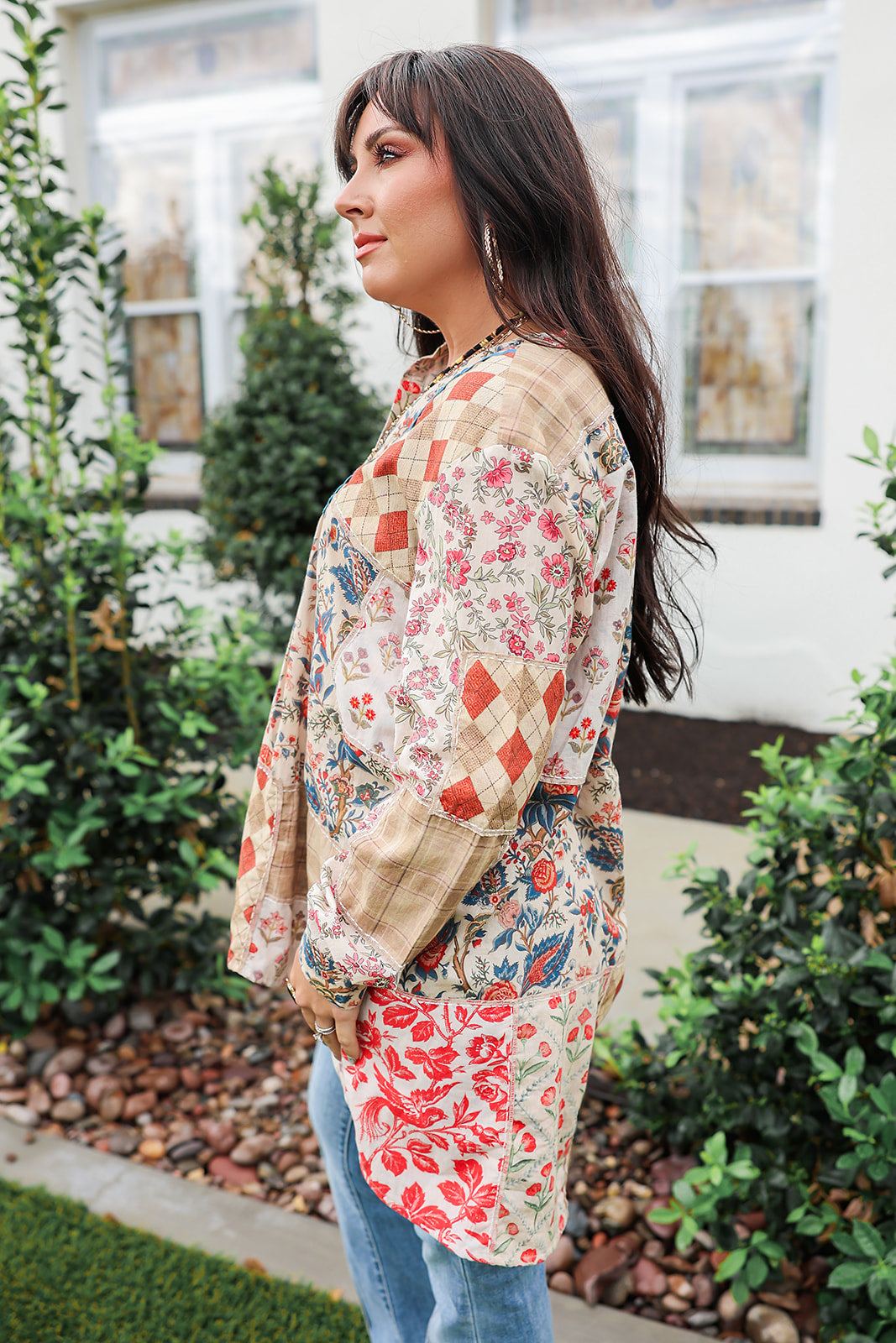 Woman with dark hair standing in a side profile position wearing the Nina Top in a lifestyle scene. The focal point of this image is the red/blue patchwork print and well as the long sleeves and length of top. The model paired this top with light-wash denim jeans.