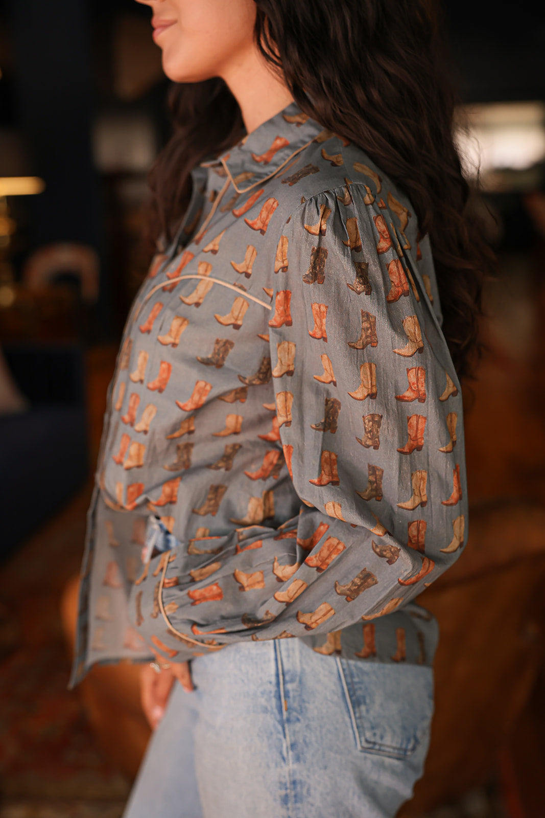 Woman with dark hair standing in a side profile position wearing the Oakley Top in a lifestyle scene. The focal point of this image is the boot graphics on sleeves and front of top, and the outlining on cuff, chest, and collar. The model paired this top with light-wash denim jeans.