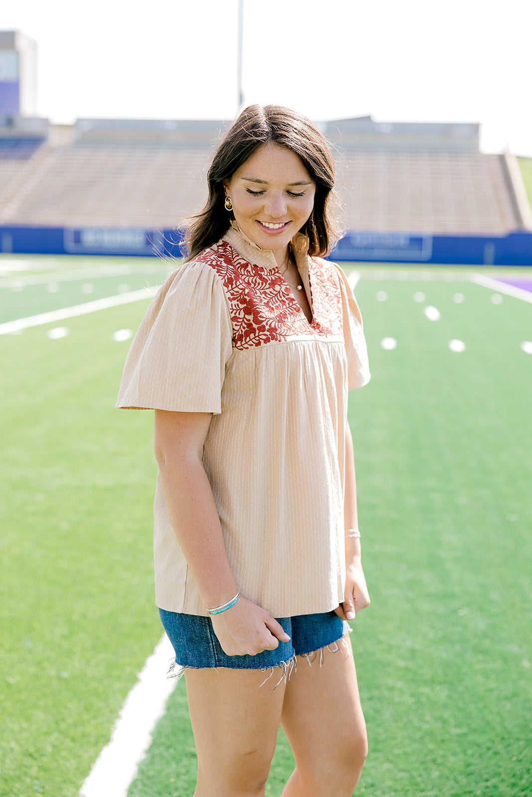 Woman standing to the side with a short-sleeve orange top featuring burnt orange embroidery, and dark-wash denim shorts.