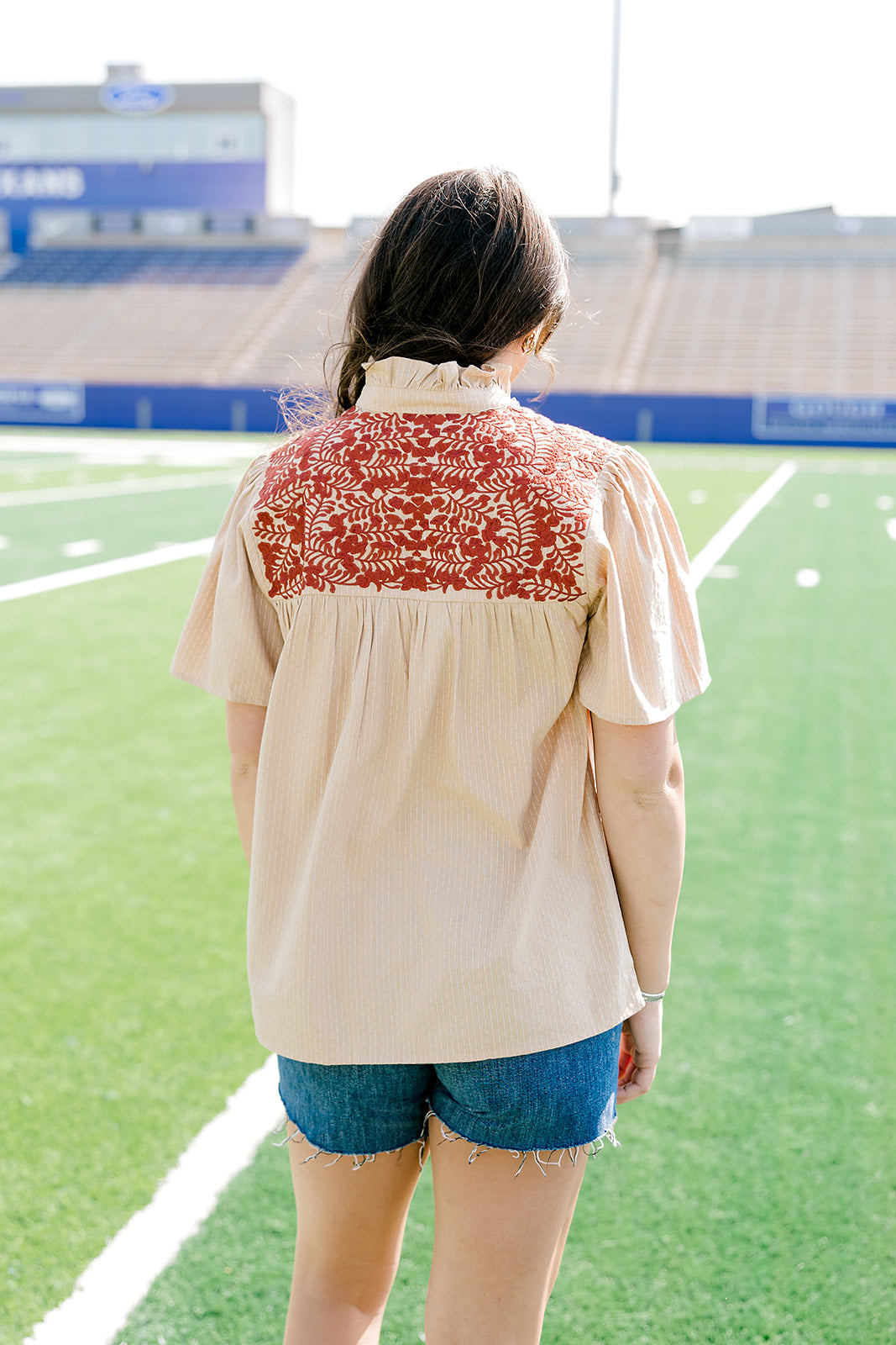 Woman with dark hair standing in a back profile position wearing a short-sleeve orange top with burnt orange embroidery, and dark-wash denims shorts.