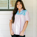 Woman with dark hair wearing a top in the Reece body style. Fabric is pink iridescent and is accented with turquoise embroidery along shoulders and chest. Model paired this top with faux leather leggings.