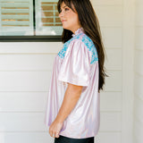 Woman with dark hair wearing a top in the Reece body style. Fabric is pink iridescent and is accented with turquoise embroidery along shoulders and chest. Model paired this top with faux leather leggings.