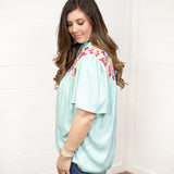 Woman with dark hair standing in a side profile position wearing the Reece Top standing against a white background. The focal point of this image is the ruffled sleeves and embroidery on top of the shoulder. The model paired this top with dark-wash denim shorts.